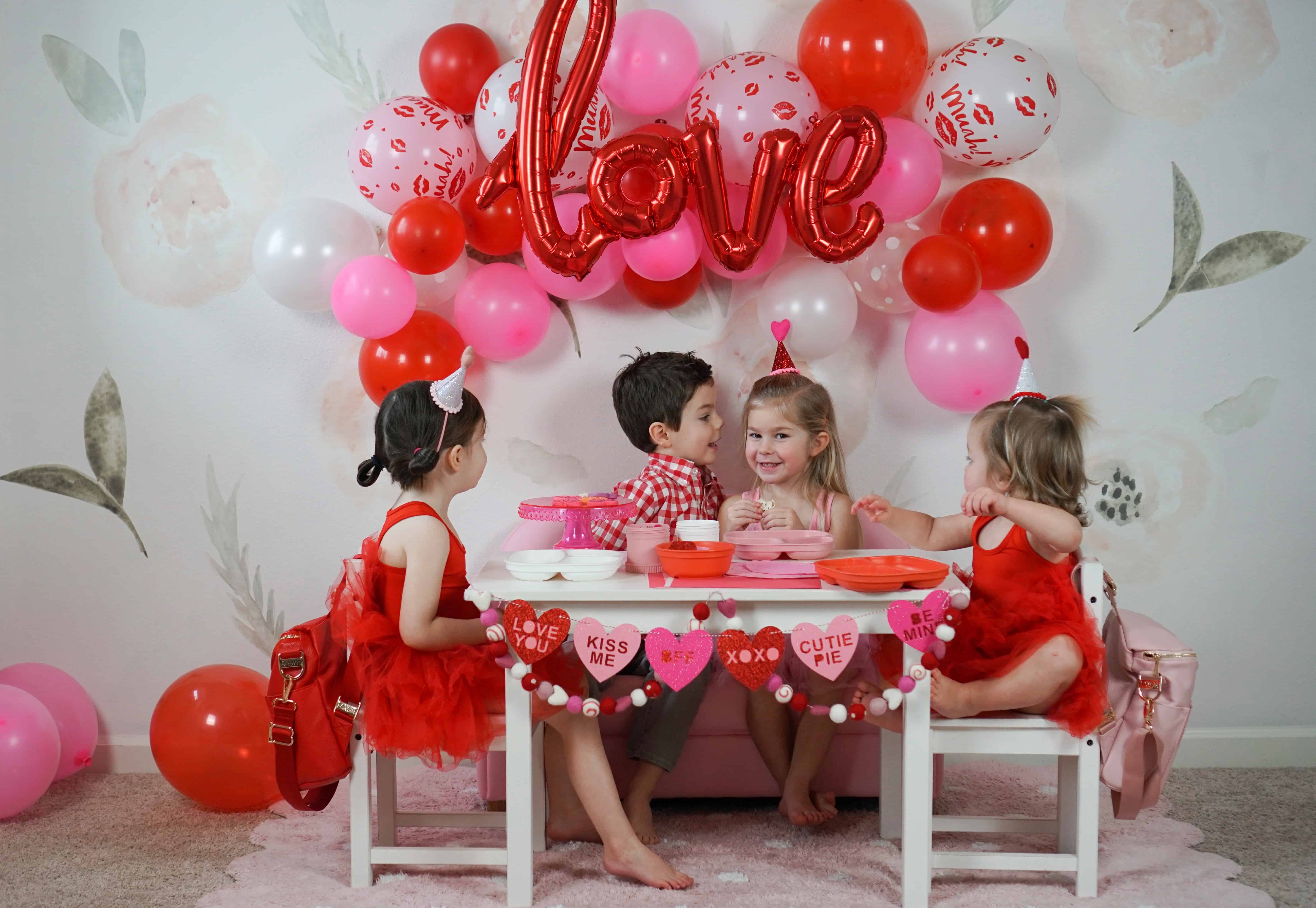 toddlers in front of pink and red balloons at white decorated table for valentines day