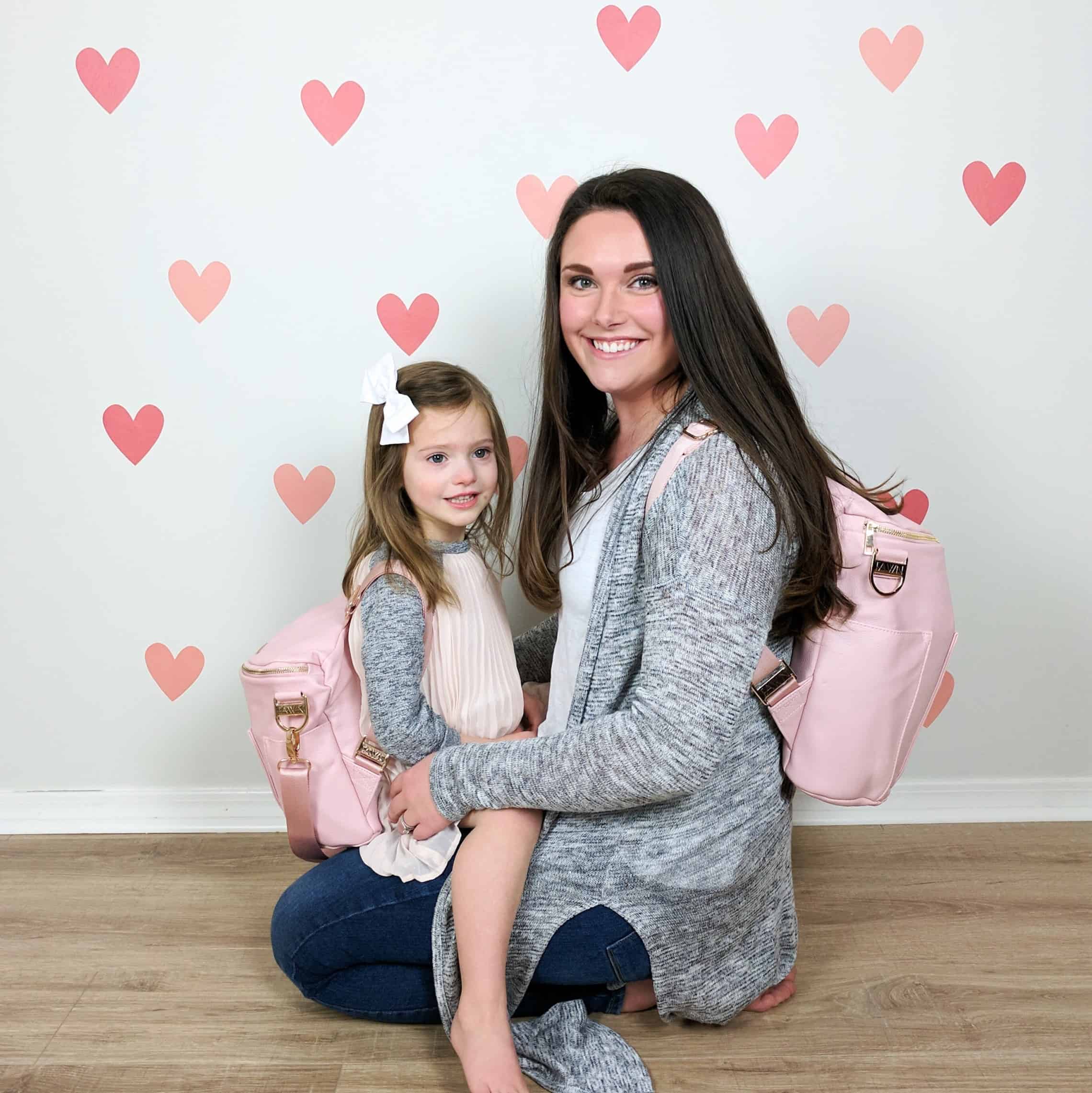 mom and daughter in matching fawn design diaper bags against pink heart wall