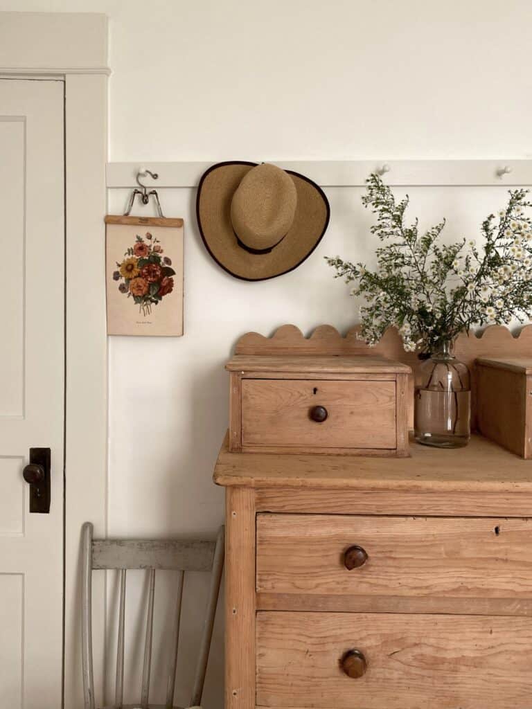 A pegboard with hanging hat, embroidered flower hanging, and a natural wooden desk with vase of wild flowers.