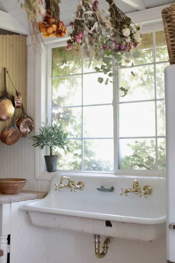 Older vintage style sink with two sets of gold faucets and flowers drying from the ceiling.