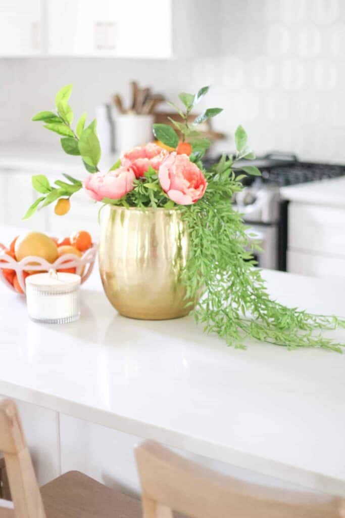 Close up of a gold vase with pink and green flowers next to a pink bowl of oranges and a candle.