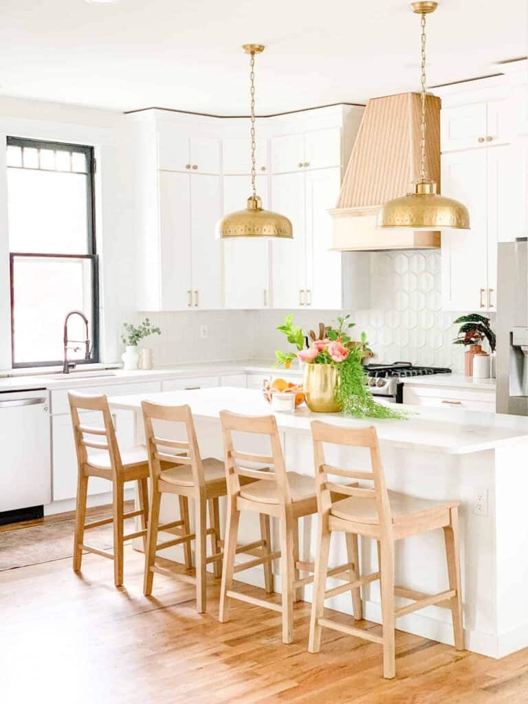 White kitchen with natural chairs and a gold vase with floral accents on a large white island.
