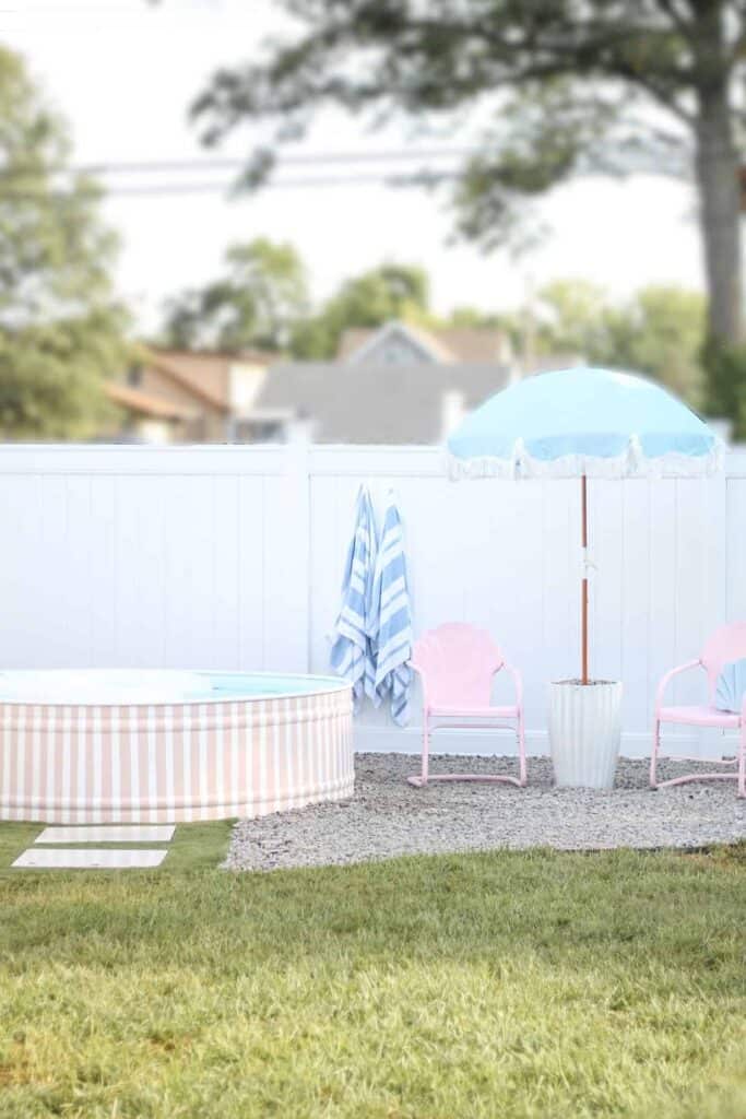 Pink and white stock tank pool with blue umbrella and pink chairs.