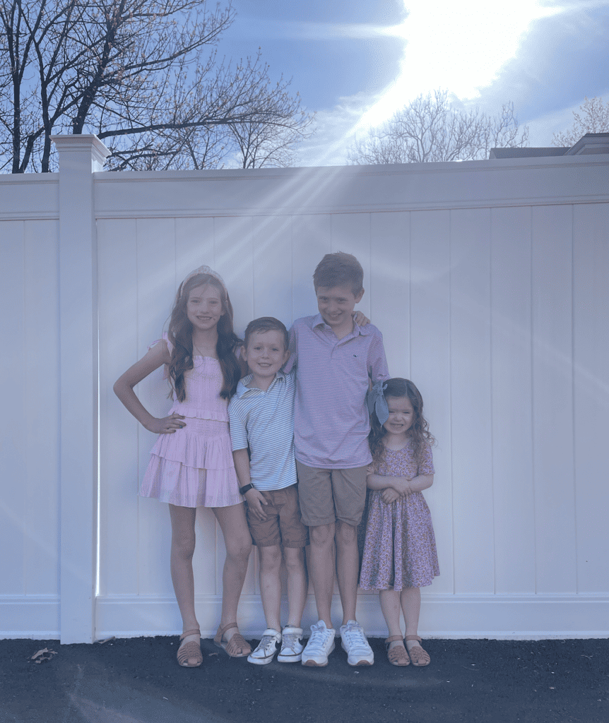 4 kids are smiling while leaning on a white fence.